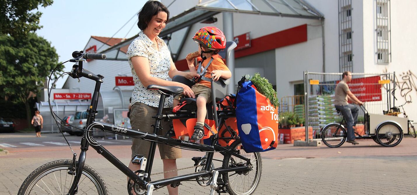 Frau schnallt Kind im Kindersitz an. Es sitzt hinten auf dem Fahrrad, dahinter sind noch Gepäcktaschen angehängt.