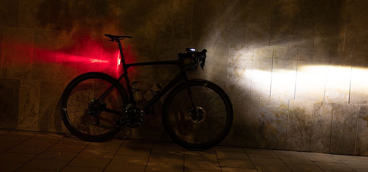 Ein Fahrrad steht in der Dunkelheit an einer Wand, mit am Lenker montiertem, eingeschalteten Scheinwerfer und an der Sattelstütze montierten, leuchtenden Rücklicht.