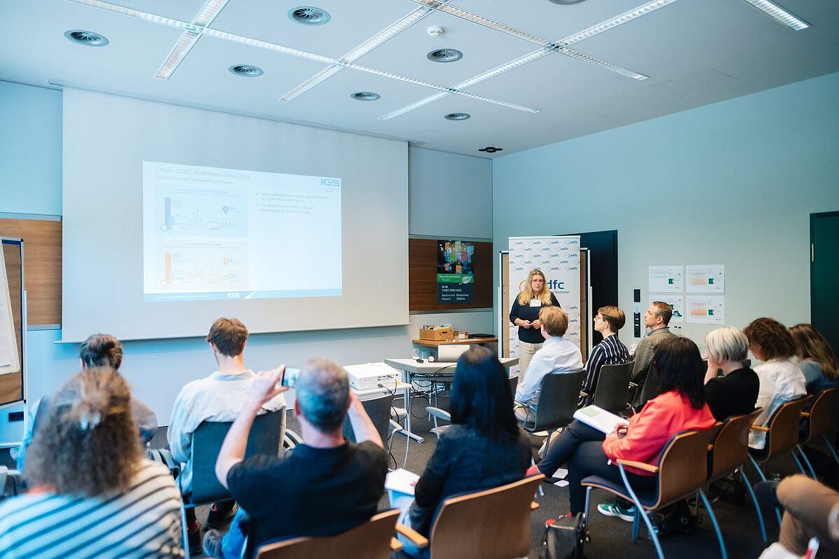 Sessions In einem Raum präsentiert eine Frau mithilfe einer Wandprojektion. Im Vordergrund ist Publikum sichtbar.
