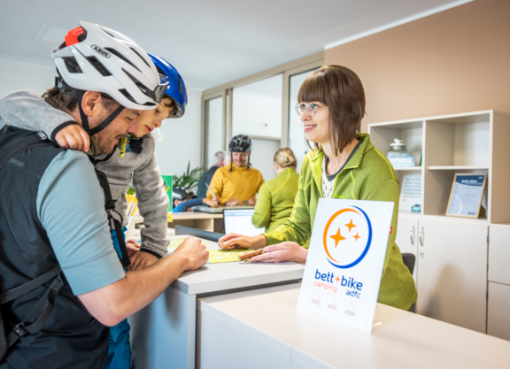 Ein Vater mit Kind auf dem Arm informiert sich an einer Rezeption eines Campingplatzes, beide tragen Fahrradhelme. Im Vordergrund steht ein Bett+Bike-Camping-Schild.
