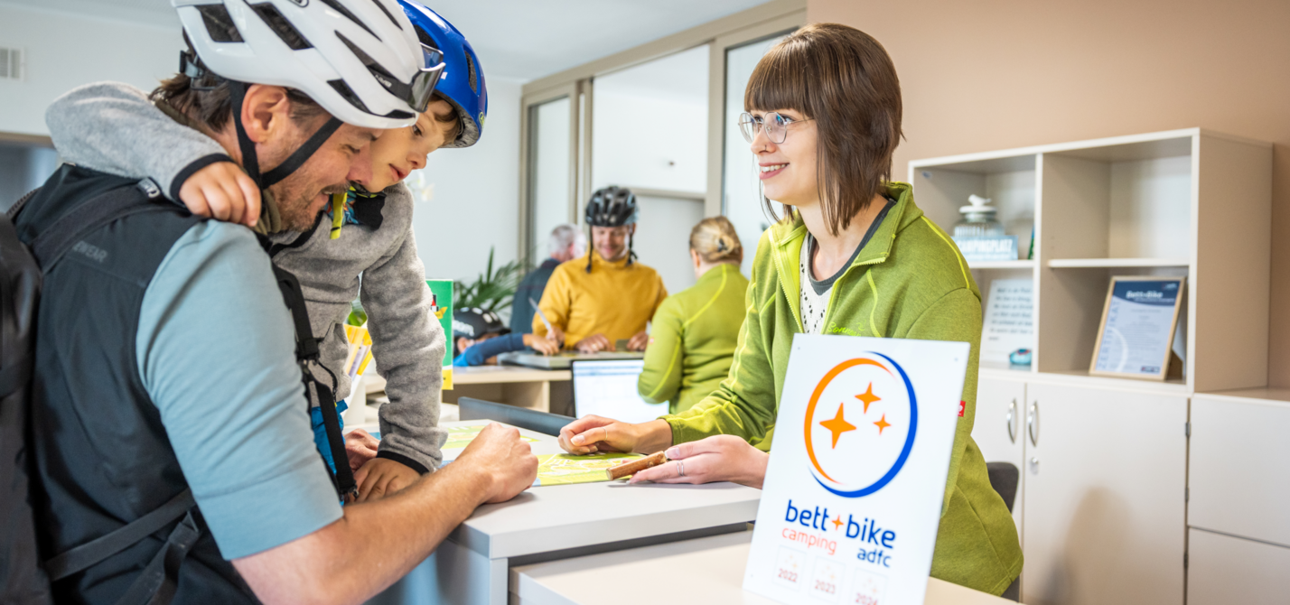 Ein Vater mit Kind auf dem Arm informiert sich an einer Rezeption eines Campingplatzes, beide tragen Fahrradhelme. Im Vordergrund steht ein Bett+Bike-Camping-Schild.