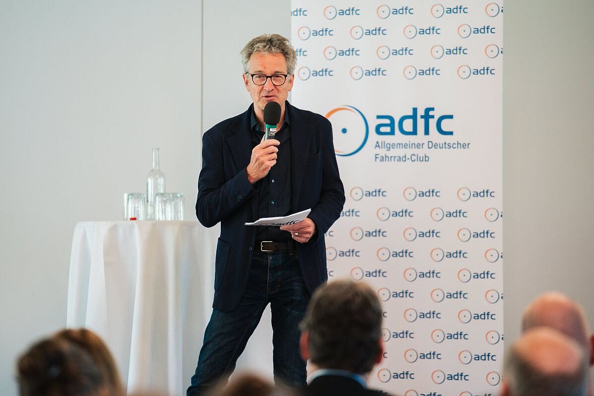 Tänzler Bühne ADFC-Bundesvorstandsmitglied Christian Tänzler steht mit Mikrofon auf der Bühne des ADFC-Radtourismuskongresses. Im Hintergund eine weiße Wand und ein weißer Aufsteller mit ADFC-Logos.