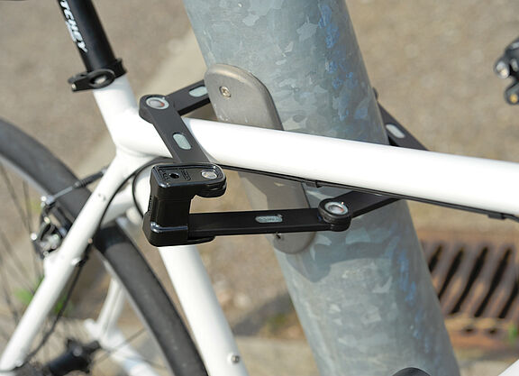 weißes Fahrrad ist mit einem Faltschloss an eine Metallstange angeschlossen