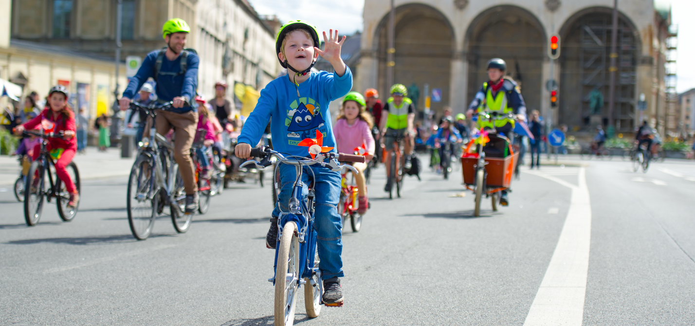 Kidical Mass Titelbild Kinder fahren mit dem Fahrrad bei einer Familien-Fahrraddemo. Ein Kind im Vordergrund winkt in die Kamera.